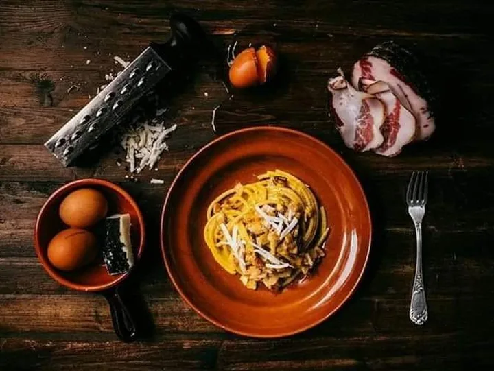 Un piatto di pasta alla carbonara in una ciotola di terracotta, su un tavolo di legno scuro, con due uova, una grattugia e un pezzo di guanciale accanto.