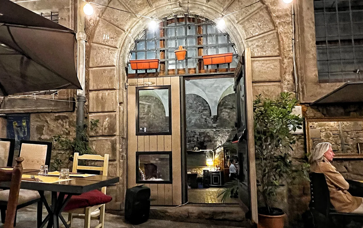 L'ingresso di MAT di sera: un portale in pietra ad arco con la porta a vetri illuminata, vasi di gerani arancioni sulla grata, una fila di lampadine tese sopra il vicolo e i tavoli apparecchiati davanti.