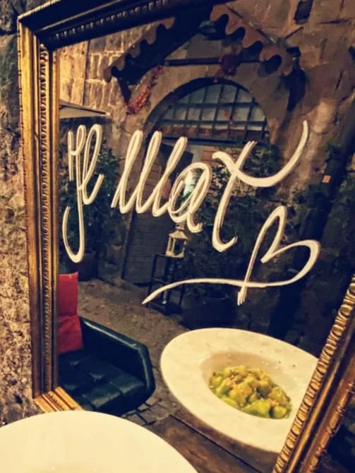 L'insegna MAT scritta a mano in luce al neon bianca su un pannello blu, riflessa dietro un piatto di pasta appoggiato al tavolo.