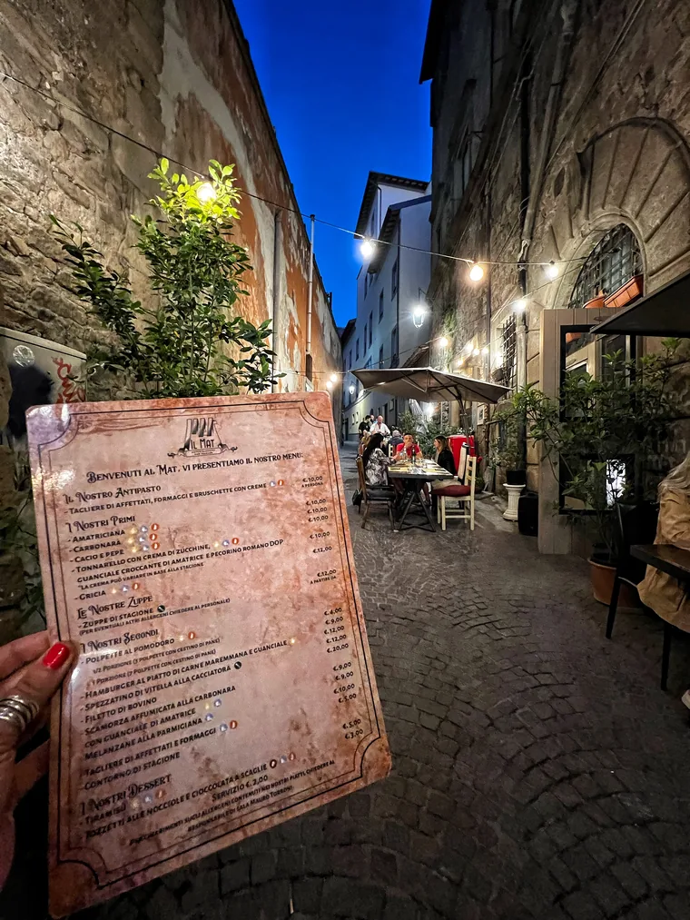 Una mano regge il menù stampato di MAT davanti al vicolo illuminato, con i tavoli occupati sullo sfondo.
