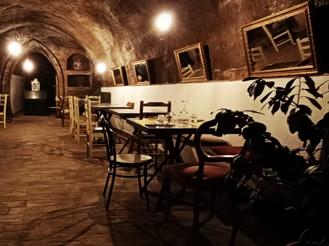 Una sala sotto la volta di pietra: tavoli apparecchiati con i bicchieri, sedie di legno diverse fra loro, cornici dorate appese al muro e lampadine a vista sotto l'arco.