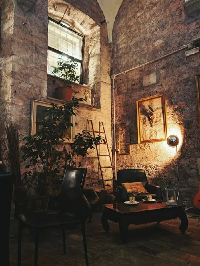 Un angolo salotto sotto la volta: due poltrone scure attorno a un tavolino basso, una finestra a grata in alto e un applique che illumina il muro di pietra.