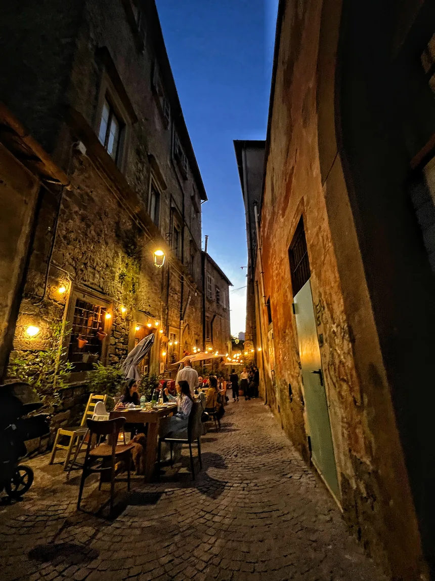 Il vicolo al tramonto: due file di tavoli di legno lungo il muro di pietra, sedie impagliate, lampadine accese sopra la strada e il cielo ancora blu in fondo.